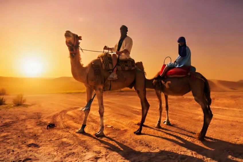 Deux personnes montent à dos de chameau dans un désert, avec un coucher de soleil en arrière-plan. 3