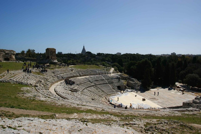 An ancient amphitheater with stone seating and a stage, surrounded by trees and ruins. 3