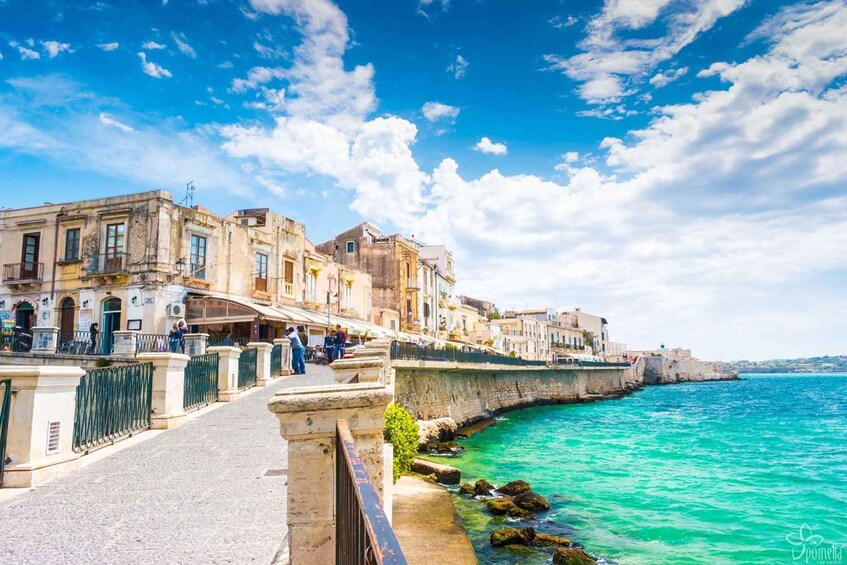 A coastal street with historic buildings, a stone wall, and turquoise sea. 4