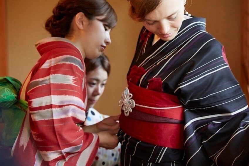 A person in a striped robe with a red sash and a brooch, being assisted by another person in a striped garment. 1