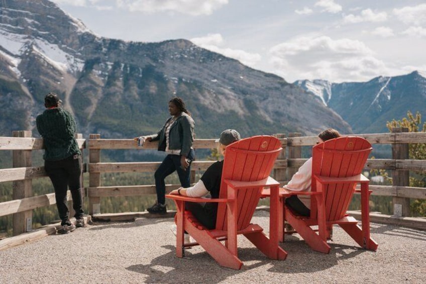 Eine Gruppe von Menschen sitzt auf roten Adirondack-Stühlen und blickt auf eine gebirgige Landschaft. 4