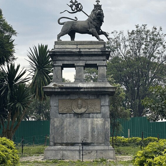 A lion statue atop a stone pedestal with a circular emblem, surrounded by greenery and a green fence. 1