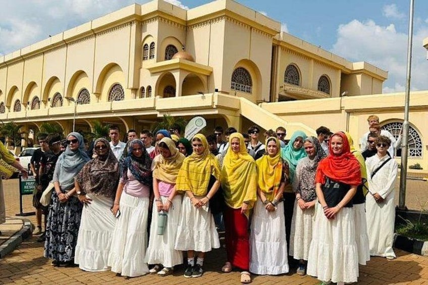 Un groupe de personnes se tient devant un bâtiment orné d’arches et surmonté d’un toit en tuiles. 1