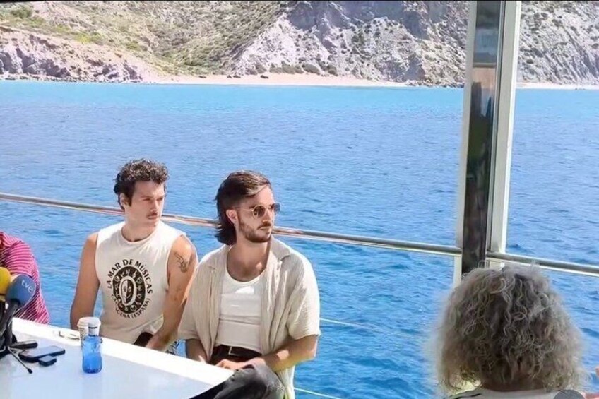 Twee mannen in een boot, met een heldere blauwe zee en rotsachtige heuvels op de achtergrond. 4