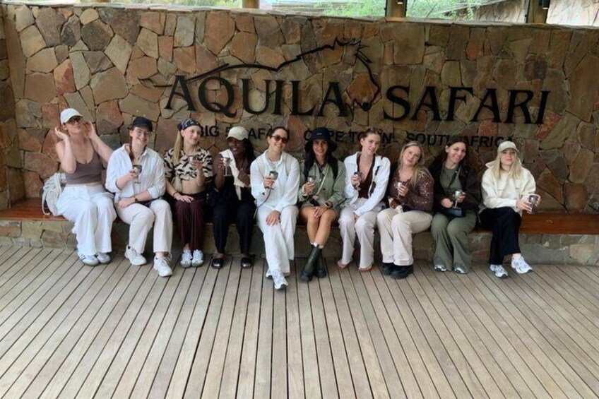En grupp människor sitter på ett trädäck framför en stenmur, och texten ”AQUILA SAFARI” syns. 1