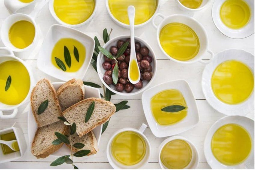 Multiple white bowls containing olive oil, a bowl of olives, and slices of bread with olive leaves. 1