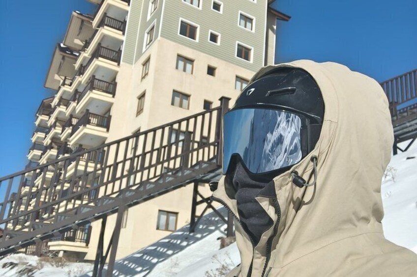 Une personne vêtue d’une veste beige et d’un casque, se tenant devant un immeuble à plusieurs étages, doté de balcons et d’un escalier en métal. 4