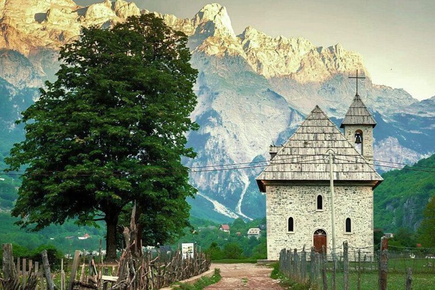 A stone church with a bell tower, surrounded by a wooden fence, a large tree, and a mountainous landscape in the background. 2