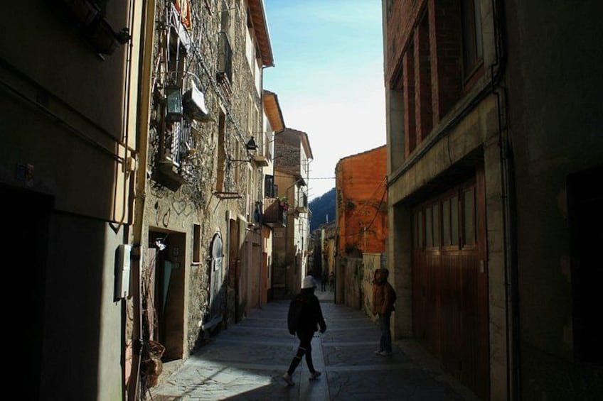 A narrow street with buildings on either side, a person walking down the center, and a statue in the distance. 5
