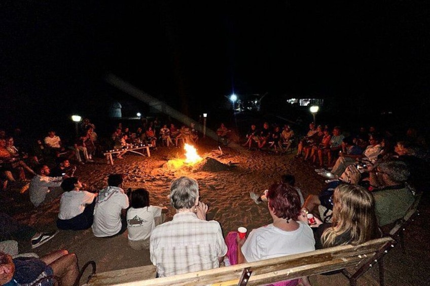 Eine Gruppe Menschen sitzt am Strand bei Nacht und beobachtet eine Feuervorführung. 4