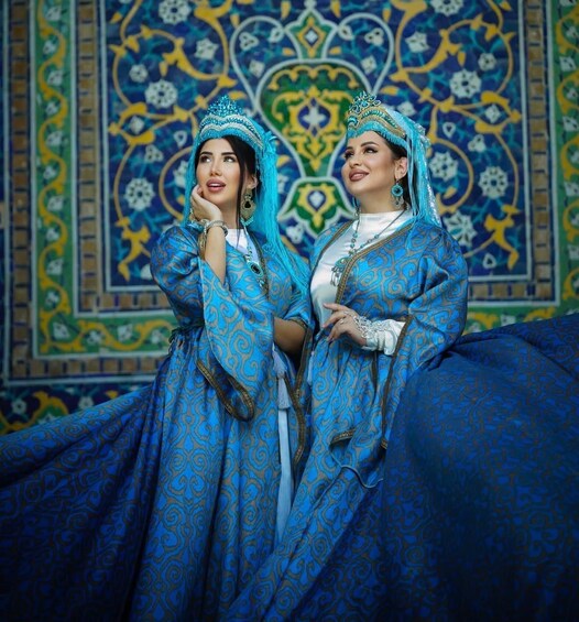 Two individuals in traditional blue attire with intricate patterns, standing in front of a detailed tile wall. 1