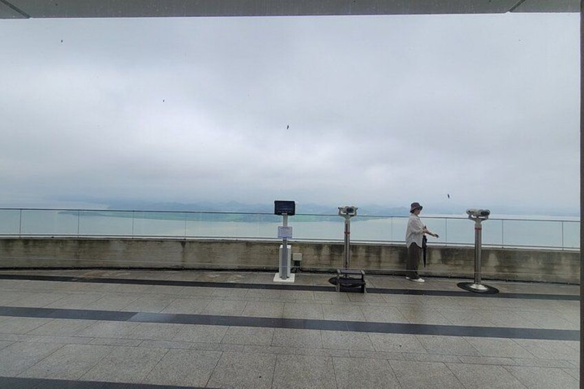 A person standing by a railing with a view of the sea, a bench, and a trash can. 3