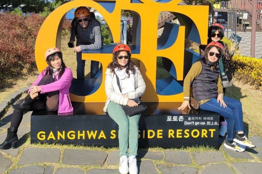 A group of people sitting in front of a large sign with the text 'Ganghwa Side Resort' and a warning message in Korean. 5