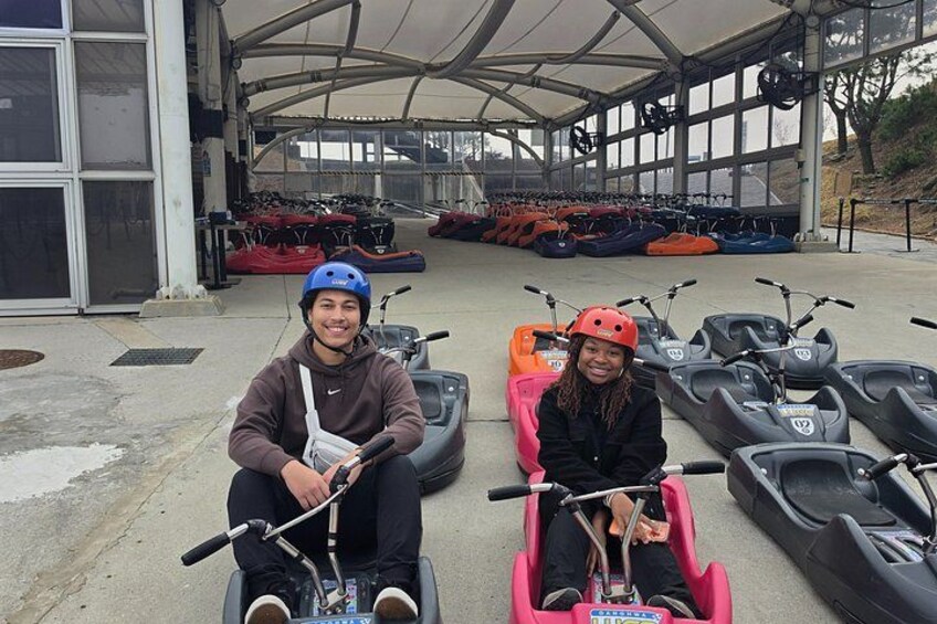Two people in go-karts with helmets, parked in a covered area with multiple go-karts lined up. 2