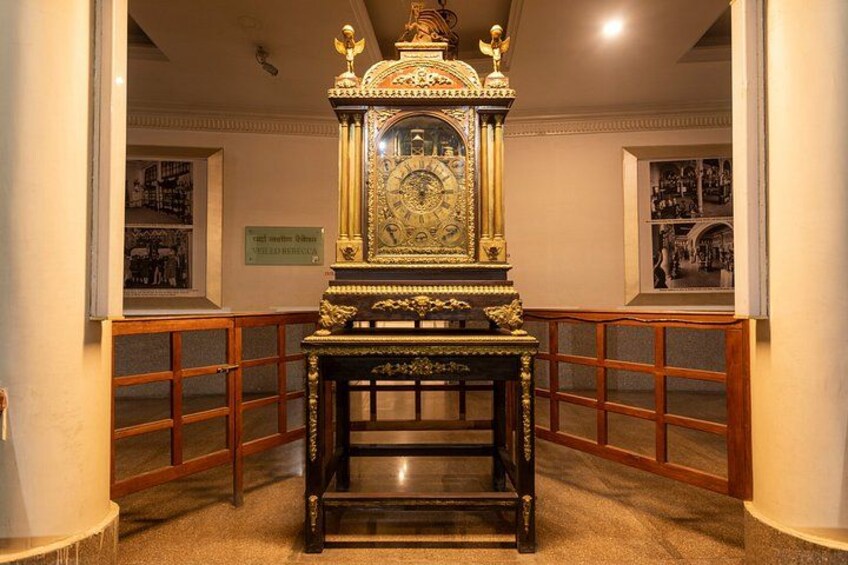Une horloge richement décorée, encadrée d’or, posée sur un piédestal en bois sombre dans un musée. 3