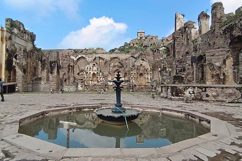 Une ruine antique avec une fontaine centrale, des murs de pierre et un ciel dégagé. 1