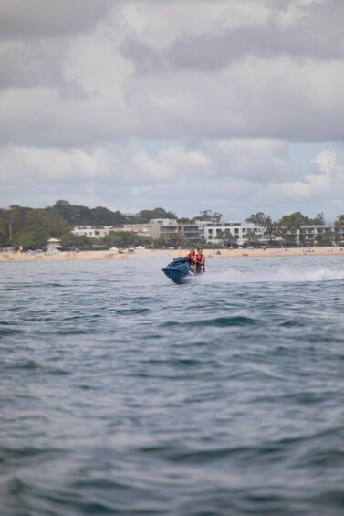 A jet ski with two riders on a body of water, with a beach and buildings in the background. 3
