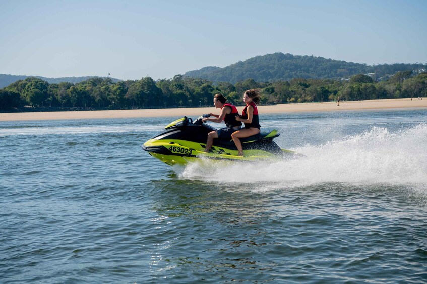 Two people on a jet ski in a body of water with a beach and hills in the background. 4