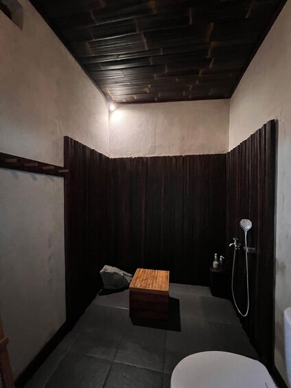 A small, dimly lit bathroom with a wooden bench, a toilet, and a shower. 3