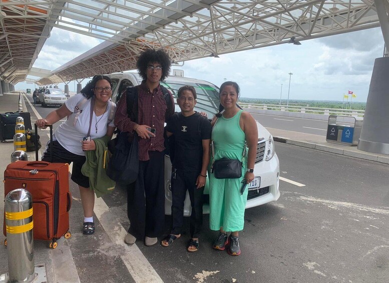 A group of people standing at a bus terminal with luggage and a white bus in the background. 1