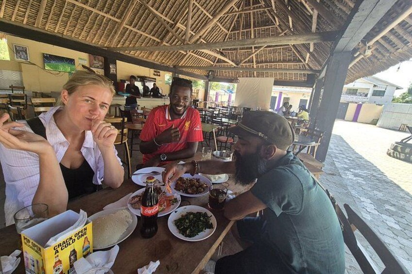 A group of people dining at an outdoor restaurant with a thatched roof, wooden tables, and various dishes on the table. 4
