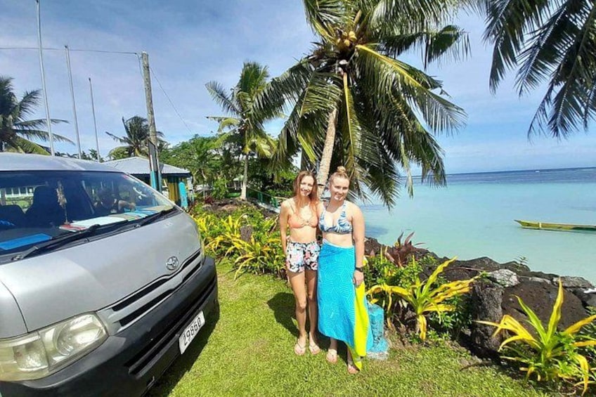 Dos personas de pie junto a un coche en una zona verde con palmeras y vista al océano. 1