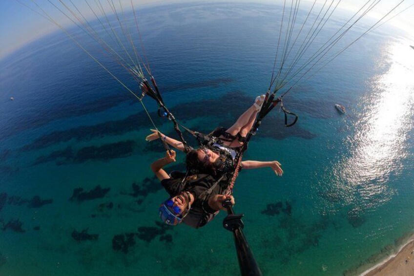 En person paraglider over et kystområde med krystalklart, blåt vand og en strand. 3
