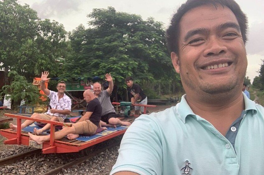 A group of people sitting on a wooden platform outdoors, with trees and a cloudy sky in the background. 3
