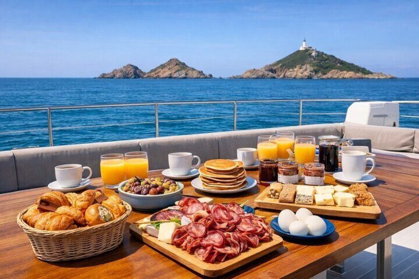 Una mesa de desayuno con croissants, huevos, embutidos y pasteles en un barco con vista al mar y un faro. 1