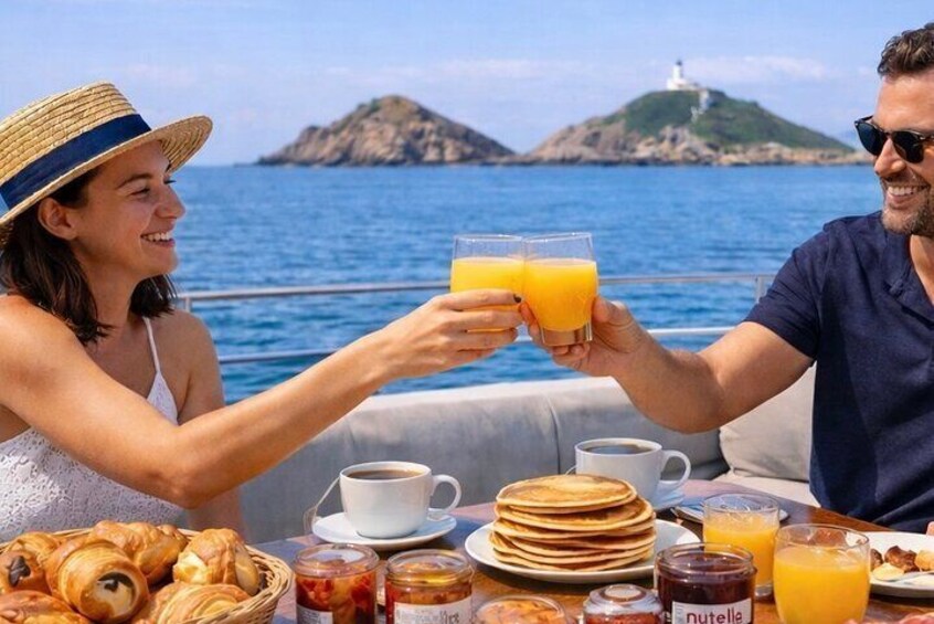 Una pareja brindando con jugo de naranja en un barco, con comida para el desayuno y una vista panorámica del mar y las islas. 5