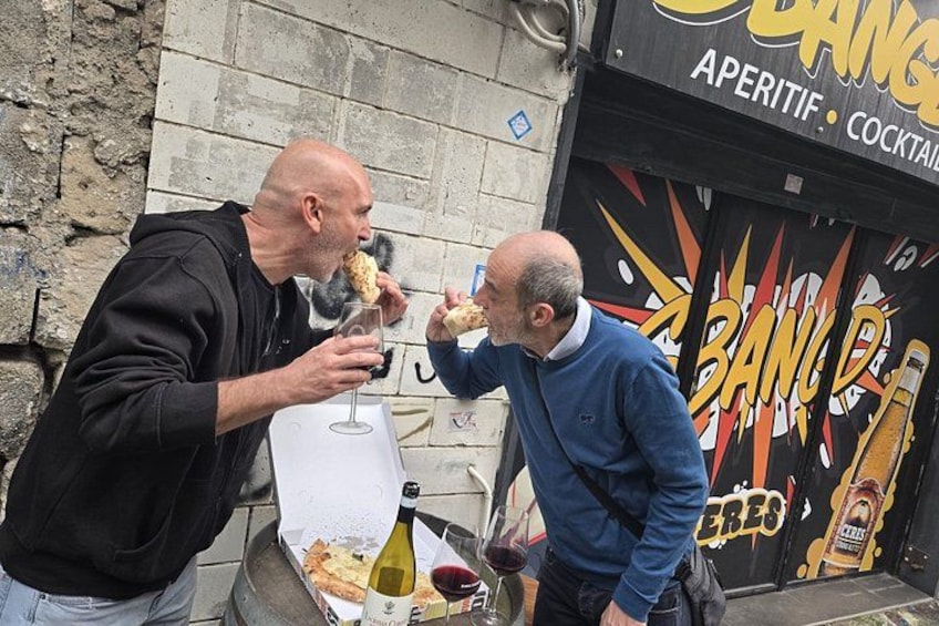 Deux hommes sont assis devant un bar, ils mangent de la pizza et boivent du vin. 3