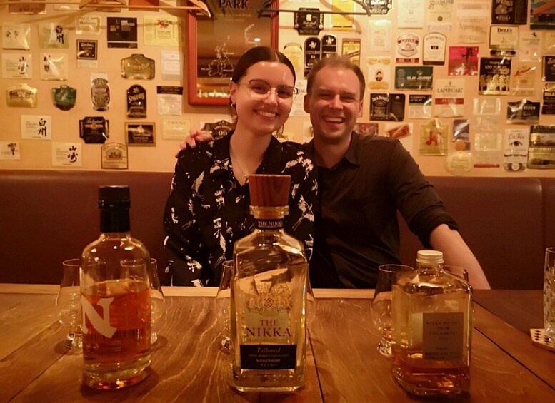 Deux personnes sont assises à une table devant des bouteilles de whisky et des verres. En arrière-plan, on aperçoit un mur décoré de diverses bouteilles et étiquettes de spiritueux. 3