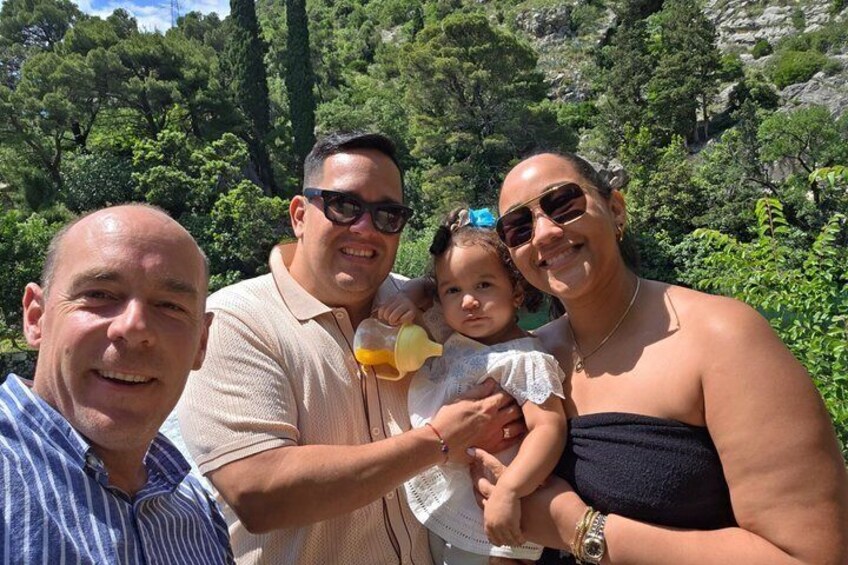 A family outdoors with a child holding a drink, surrounded by greenery and trees. 2