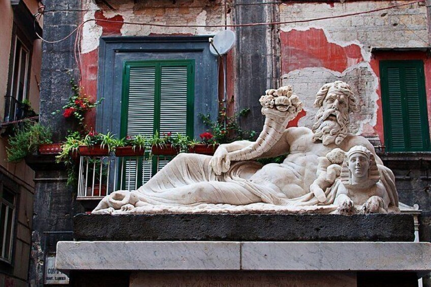 A marble statue of a reclining man holding a child, set against a building facade with green shutters and potted plants. 3