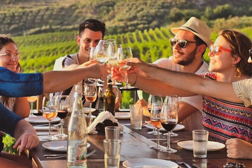 A group of people toasting with wine glasses at a table with bottles and glasses of wine, set against a vineyard backdrop. 1