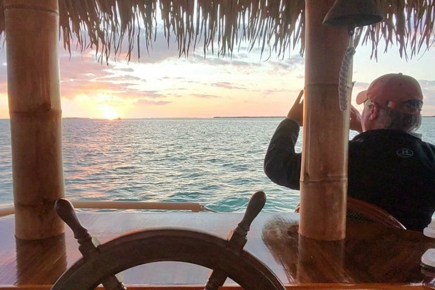 Eine Person steuert ein Boot mit einem Steuer, Palmen und einen Sonnenuntergang über dem Meer. 4
