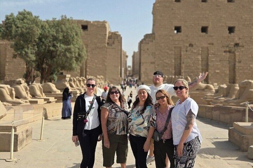 A group of people standing in front of ancient stone structures, possibly ruins, with trees and a clear sky in the background. 3