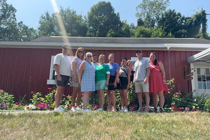 A group of people standing in front of a red building with a white door and windows, surrounded by grass and flowers. 2