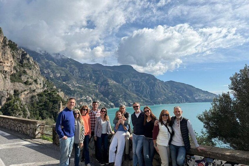 Un groupe de personnes se tient sur une route côtière, avec une chaîne de montagnes en arrière-plan et une étendue d’eau. 5