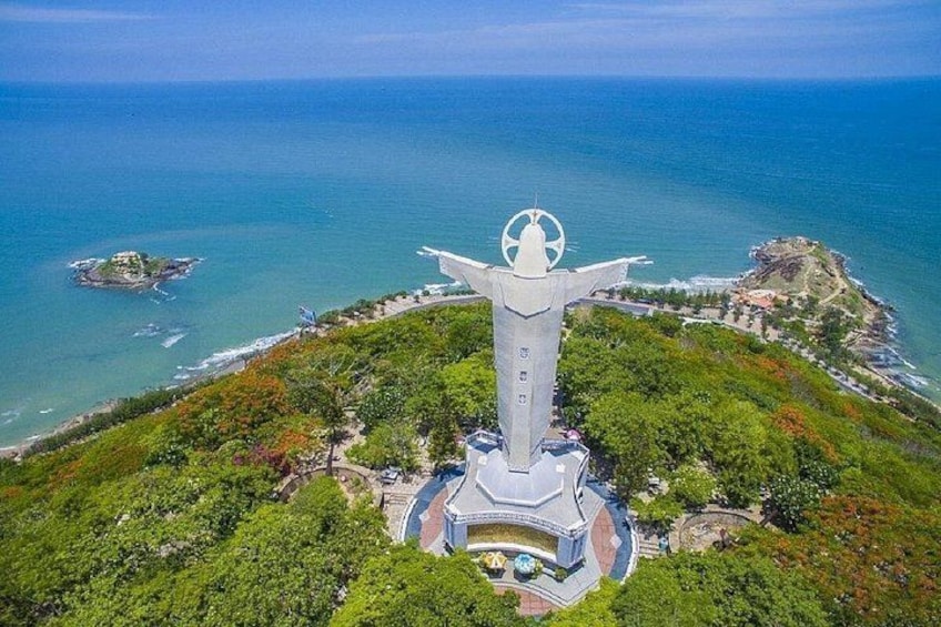 Eine große Statue mit ausgestreckten Armen, umgeben von üppiger Vegetation und mit Blick auf eine Küstenlinie. 2