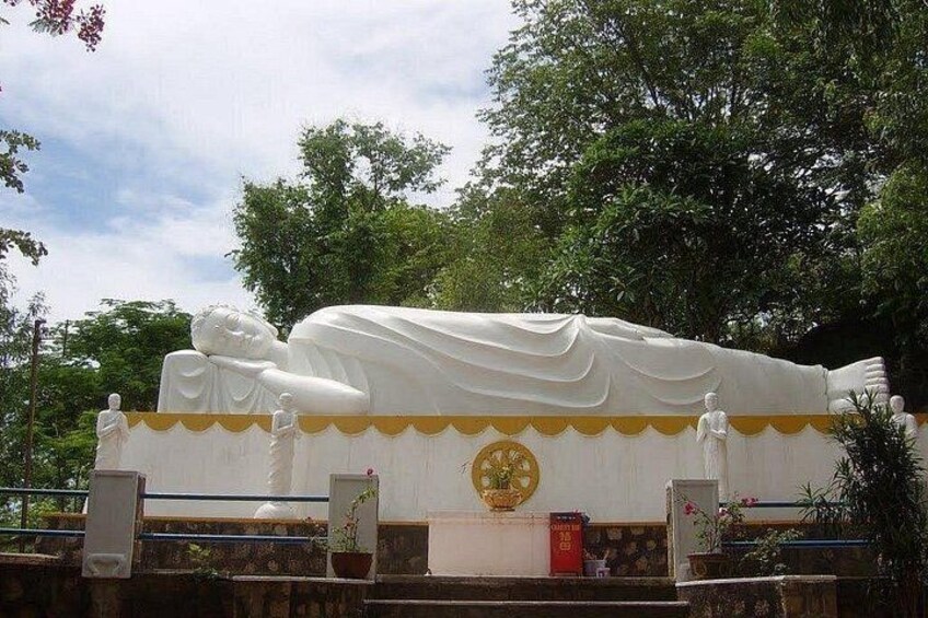 Eine große weiße Statue einer liegenden Figur, möglicherweise ein religiöses Symbol, mit einem goldenen Emblem auf der Brust, umgeben von kleineren Statuen und einer dekorativen Bordüre. 4