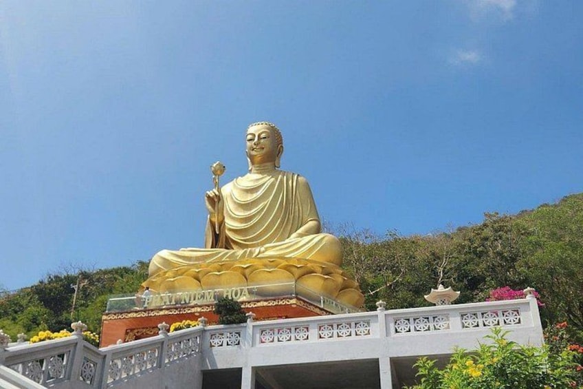 Eine große goldene Buddha-Statue sitzt auf einem Sockel, umgeben von Grün und bewacht durch ein weißes Geländer, unter blauem Himmel. 3