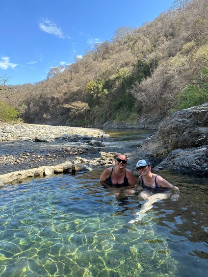 Two people in a clear river surrounded by rocky terrain and sparse vegetation. 1