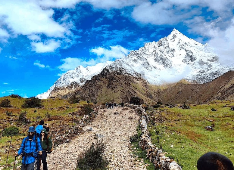 A mountainous landscape with a clear sky, a rocky path leading to a group of hikers, and a snow-capped peak in the background. 2