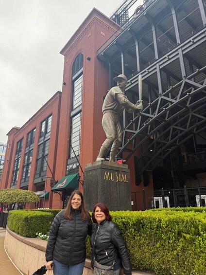 A statue of a baseball player on a pedestal labeled 'MUSIAL' in front of a brick building with large windows. 3