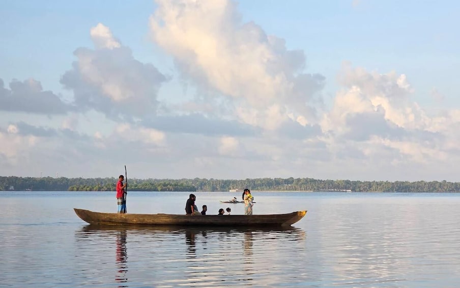 A wooden boat on calm water with people on board, a person standing on the bow, and a forested shoreline in the background. 5
