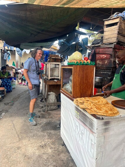 A street market with a vendor preparing food, a woman in a patterned shirt, and various goods displayed. 4