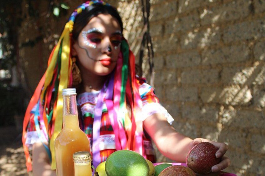 Eine Person mit farbenfroher Kleidung und Kopftuch, die Obst hält und an einem Tisch mit einer Flasche und einem Glas sitzt. 3