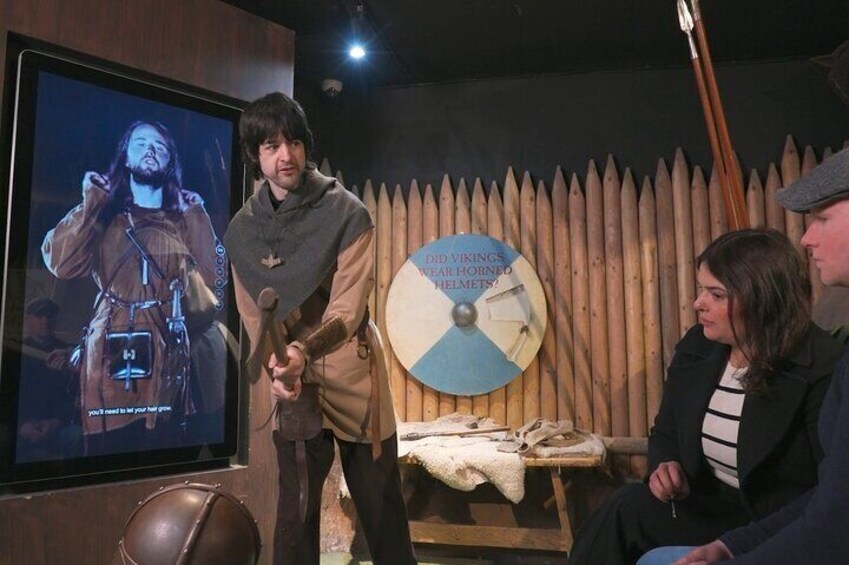 Una persona de pie frente a una pantalla que muestra un personaje con armadura, con una valla de madera y un escudo al fondo. 2