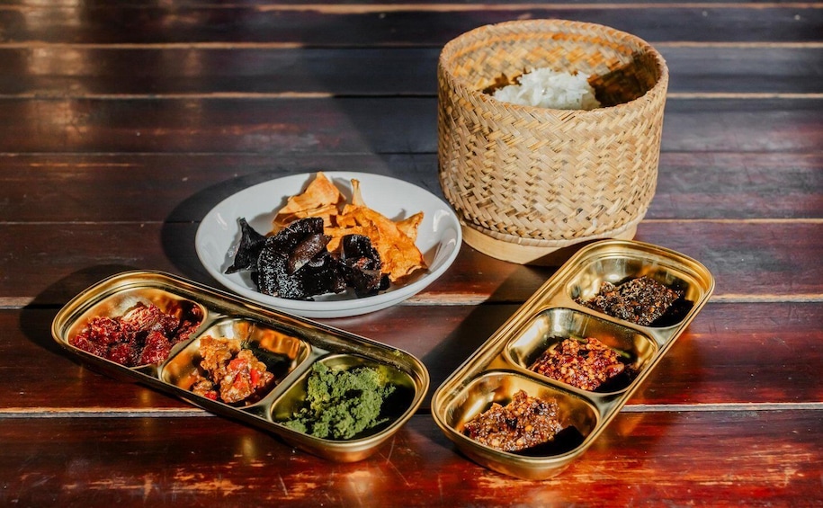 A wooden table with a woven basket, a plate of dried fish, and metal containers with various condiments. 4